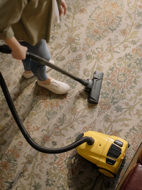 A person kneeling on a patterned carpet in a room illuminated by warm lighting, using a yellow portable vacuum cleaner with a black hose to perform surface cleaning. The individual, dressed in a beige